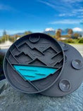 Load image into Gallery viewer, Black and blue object on a rock with a blue sky and mountains in the background
